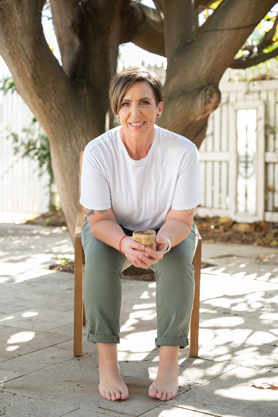 Anita Gibbons sitting barefoot under an olive tree, smiling, holding a handmade pottery mug
