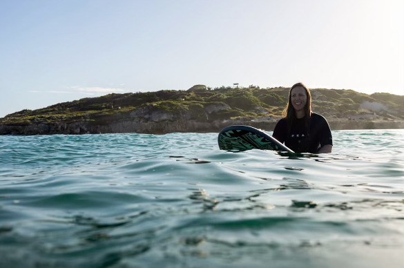 Anita Gibbons sitting calmly on a surfboard in golden evening light